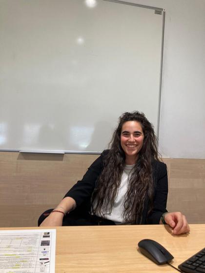 A smiling woman sitting at a desk in a classroom with a whiteboard in the background.