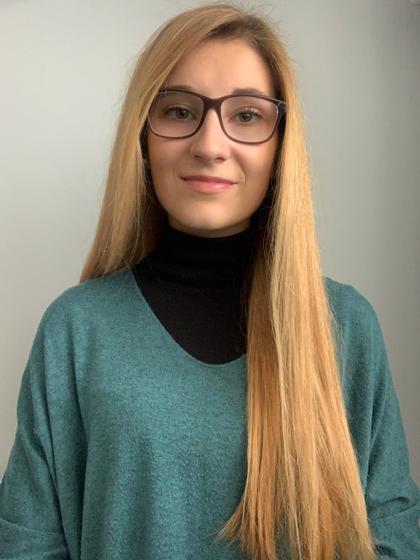 Portrait of a young woman with long blonde hair wearing glasses and a green sweater over a black turtleneck.