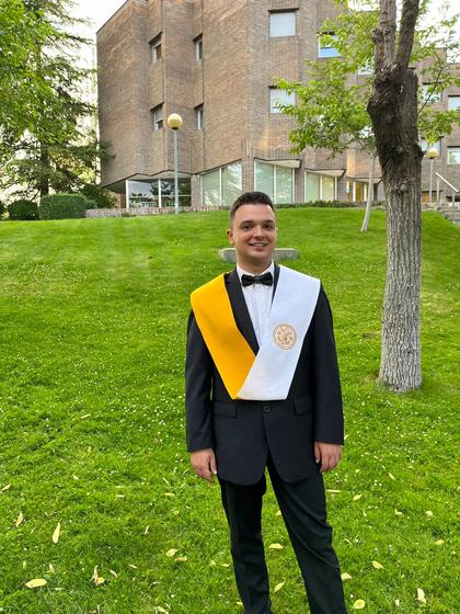 A young man in graduation attire stands smiling on a grassy area in front of a brick building.