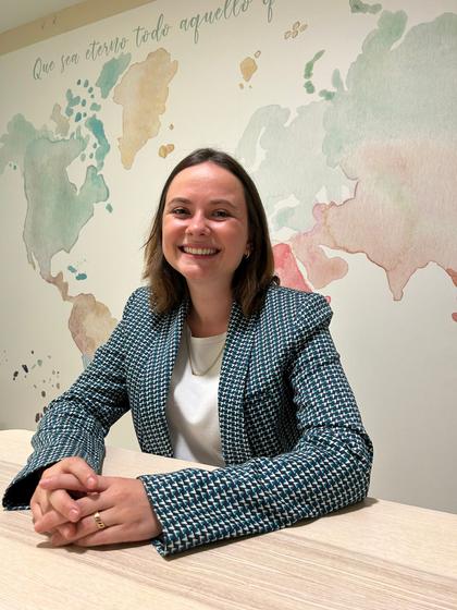 A smiling woman in a business suit sitting at a desk with a world map on the wall behind her.