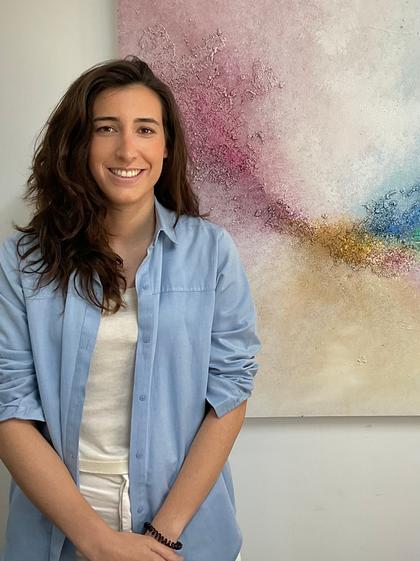 A smiling woman in a blue shirt standing in front of a colorful abstract painting.