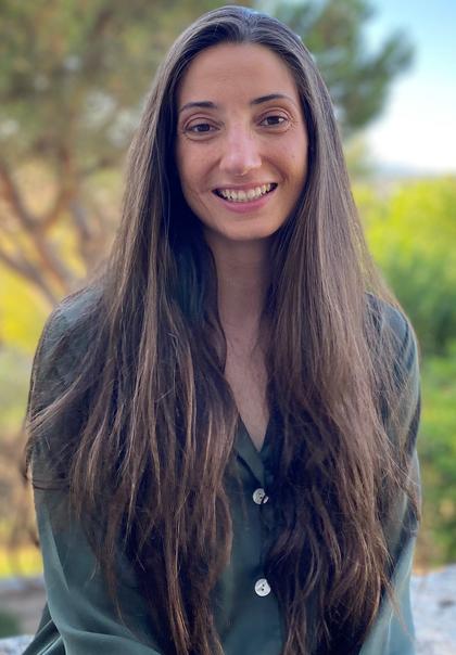 A portrait of a smiling woman with long hair, wearing a green shirt, standing outdoors.