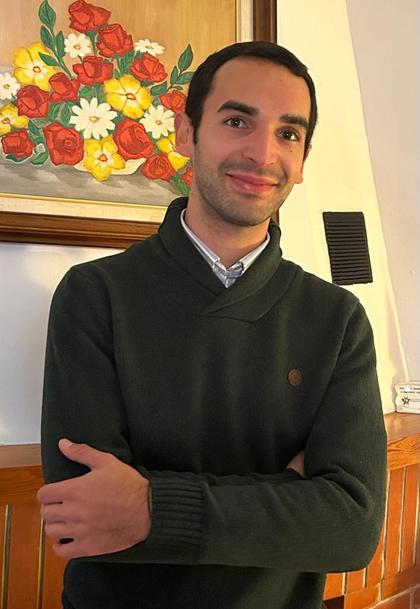 Un hombre sonriente con un suéter oscuro posa con los brazos cruzados frente a un cuadro de flores coloridas.