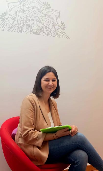 A woman sits smiling in a red chair, holding a green folder against a wall decorated with a white mandala design.