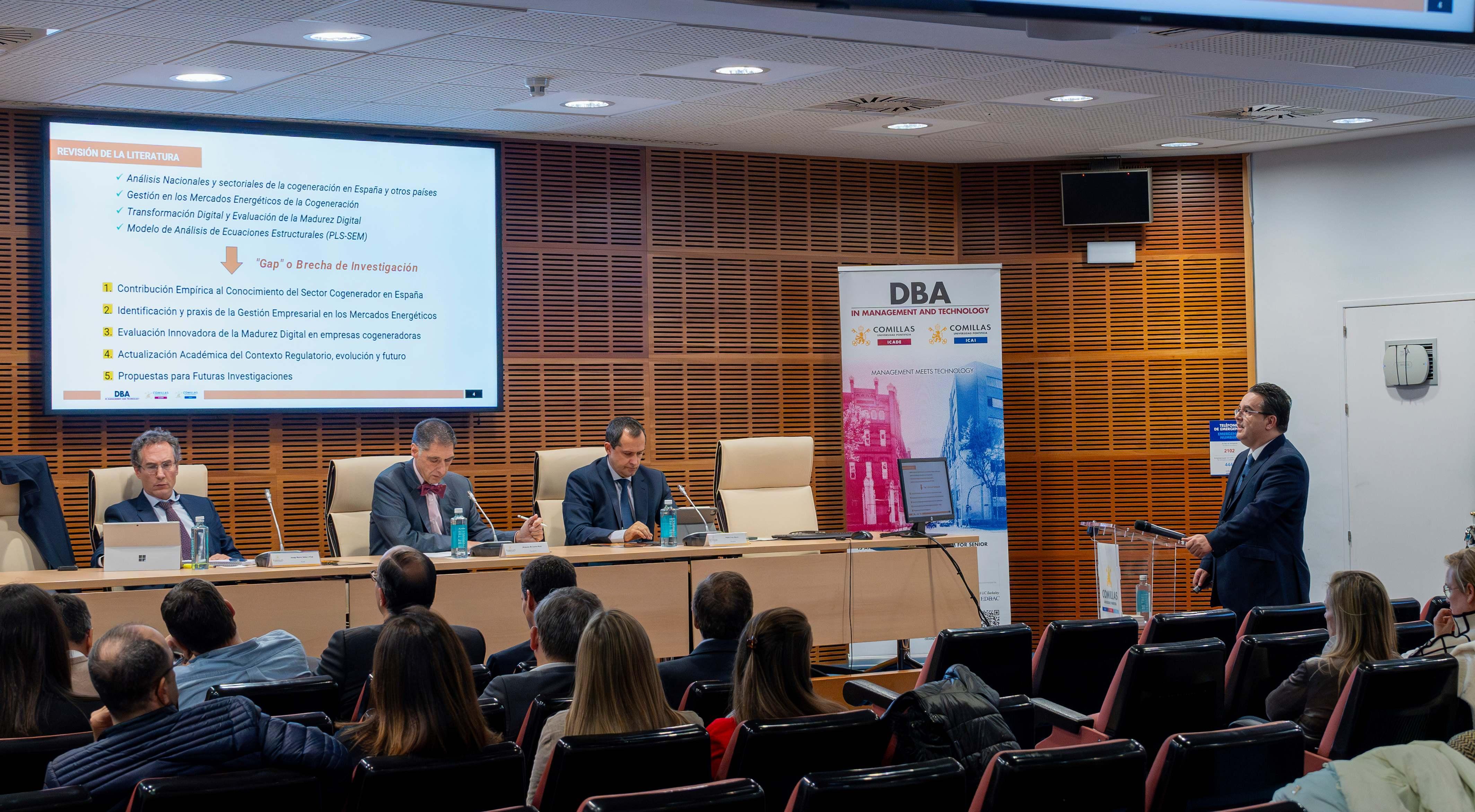 Un hombre de traje está dando una presentación en una conferencia con cuatro panelistas sentados detrás de él y una audiencia frente a él.