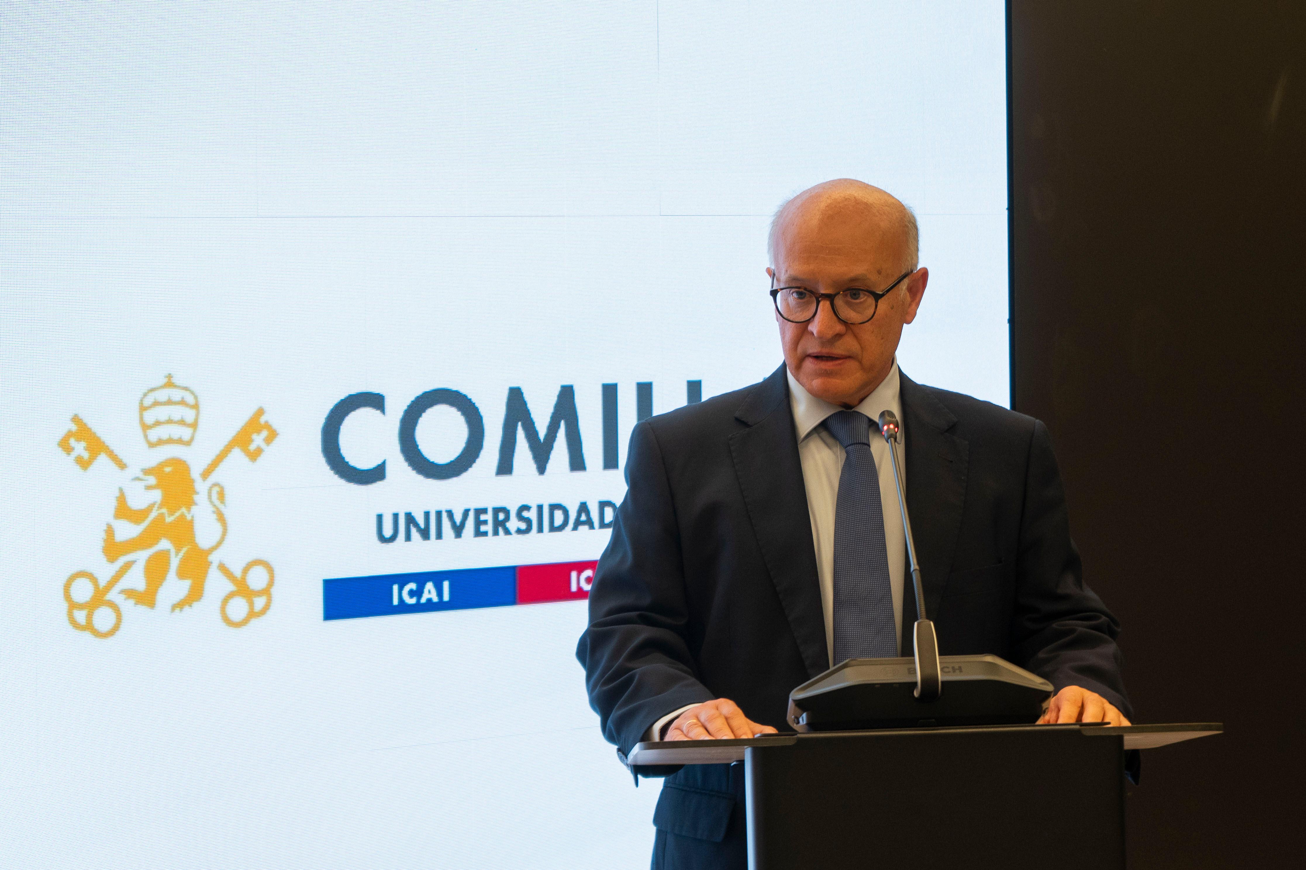 Un hombre de cabello corto y gafas se encuentra de pie frente a un micrófono, hablando en un evento relacionado con la universidad.