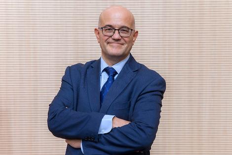 A confident bald man in a blue suit and glasses, standing with arms crossed in front of a textured beige background.