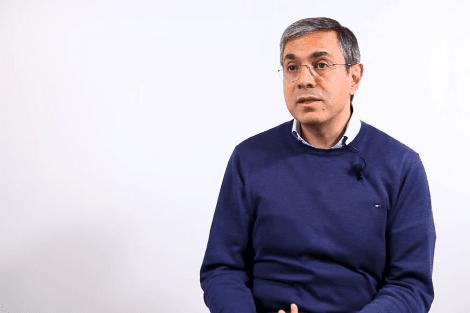 A man in a blue sweater sitting and talking against a white background.