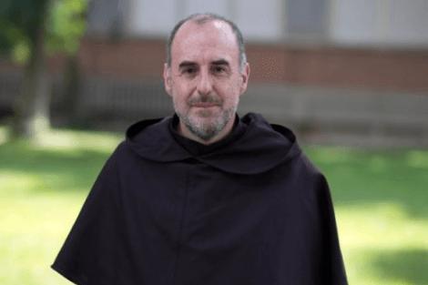 A man in a religious robe stands outdoors with trees and a building in the background.