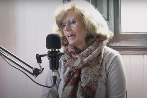 A woman with glasses and a scarf speaks into a microphone in a well-lit room.