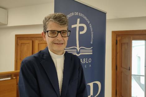 A man in a blue suit standing in front of a 'Paulo VI' banner inside a room.