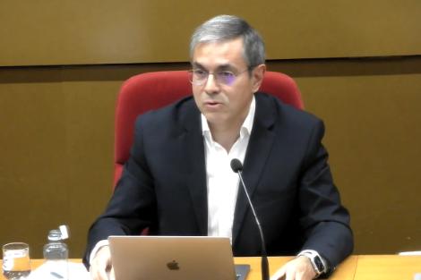 A man in a suit sitting at a desk with a laptop and microphone in a conference setting.