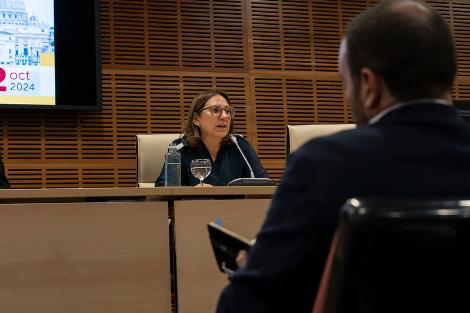 A woman speaks during a conference as a man listens from the audience in a modern hall.