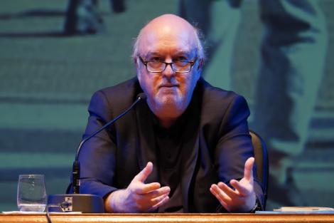 A man wearing glasses gestures while speaking at a lectern against the backdrop of a projected image.