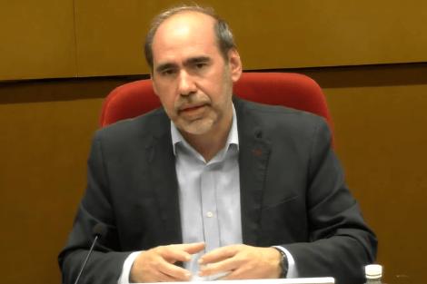 A man in a suit sits at a table with a microphone, appearing to speak in a formal setting.