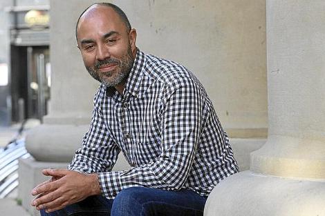 Un hombre sonriente con barba, sentado en una estructura de piedra, vistiendo una camisa de cuadros.