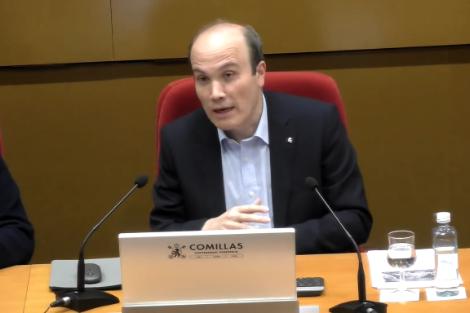 Un hombre calvo vestido con traje oscuro habla en un panel de conferencias con el logo de Comillas en el frente de la mesa.