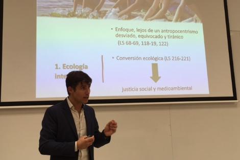 Un hombre joven está dando una presentación en una sala de clases frente a una pantalla que muestra diapositivas sobre ecología integral.