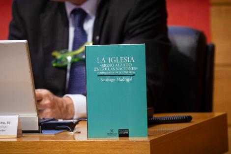 Un hombre con traje sentado detrás de una mesa en una conferencia, con un libro titulado 'La Iglesia en Tiempos Azarosos: Testimonios y discernimiento' de Santiago Madrigal visible en primer plano.