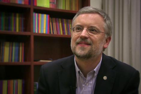 A middle-aged man with a beard, wearing glasses and a suit, seated in front of a bookshelf filled with colorful books.