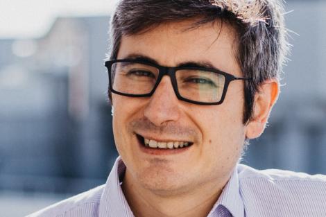 A close-up portrait of a smiling man wearing glasses and a purple striped shirt.