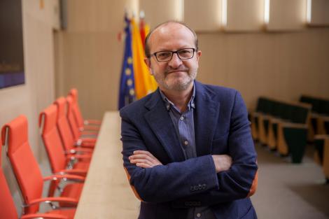 A man in a blue blazer and glasses stands with crossed arms in a conference room with red chairs and flags in the background.