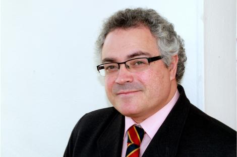 A middle-aged man with gray hair and glasses, wearing a black coat and a colorful striped tie, smiling at the camera.