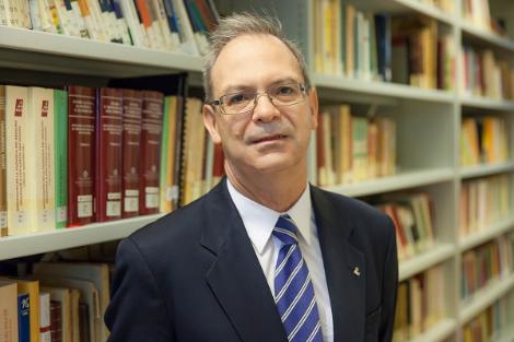 A middle-aged man wearing a blue suit and tie stands in front of a bookshelf filled with books.