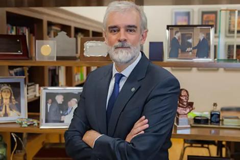 A man in a suit stands with crossed arms in an office surrounded by pictures and books.