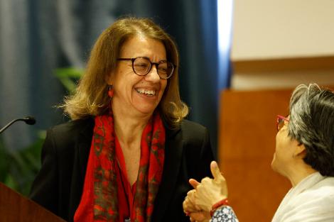 Una mujer sonriente con bufanda roja está de pie al lado de otra mujer que parece estar hablando desde un podio.