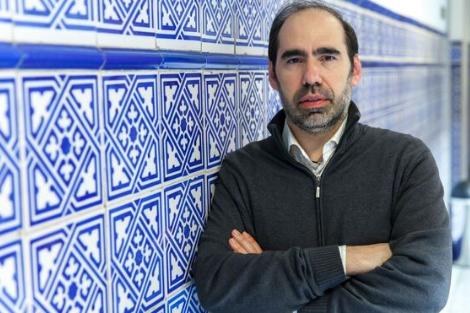 A man in a grey sweater and white shirt standing in front of a blue and white tiled wall, arms crossed.