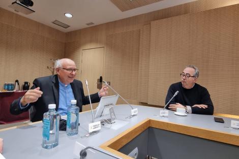 Two men engaging in a discussion at a modern conference table with microphones and bottles of water.