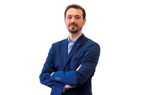 Hombre con barba, vestido con traje azul y camisa blanca, sonriendo y con los brazos cruzados.