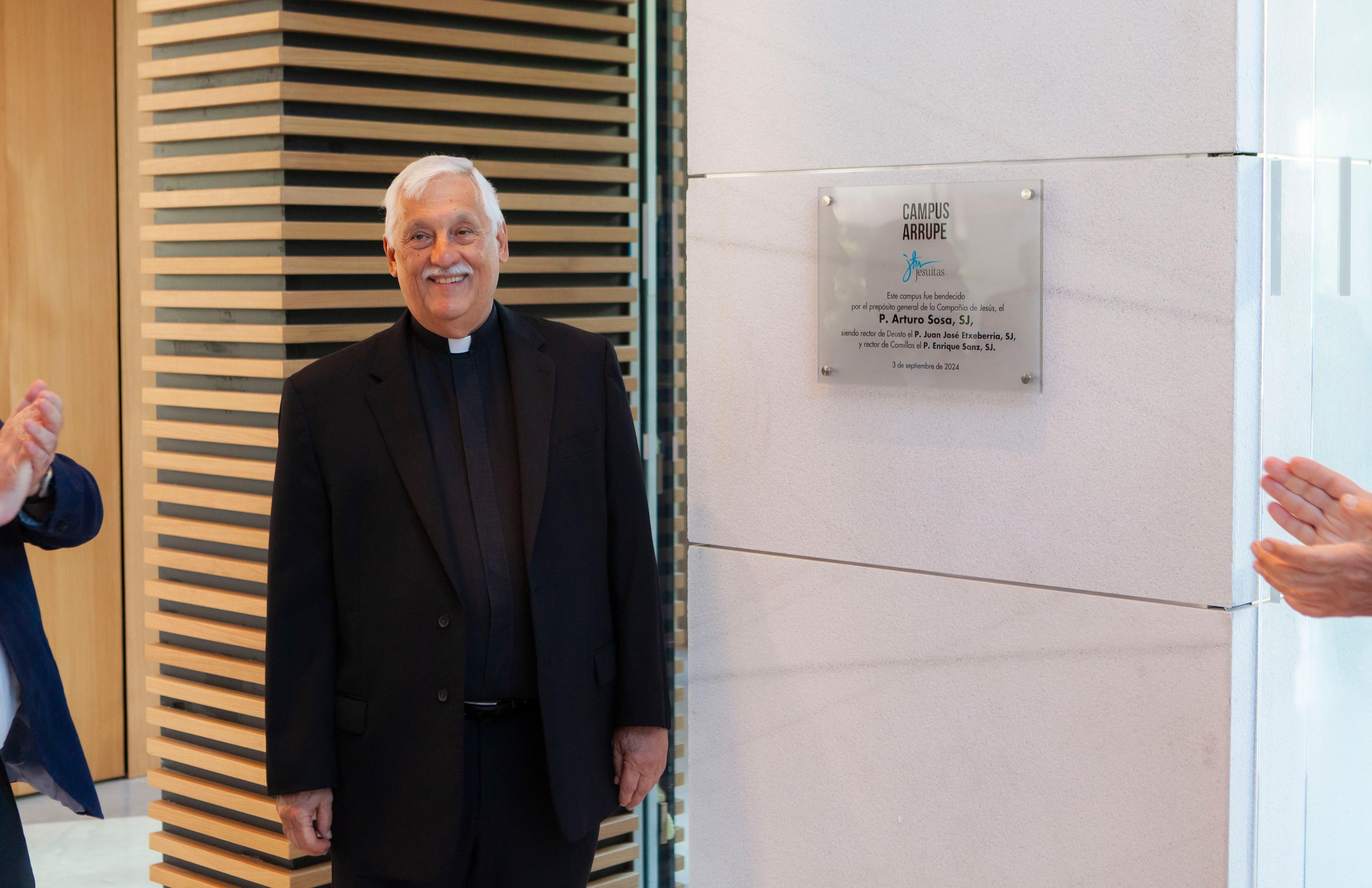 A priest smiling in front of a commemorative plaque inside a building.