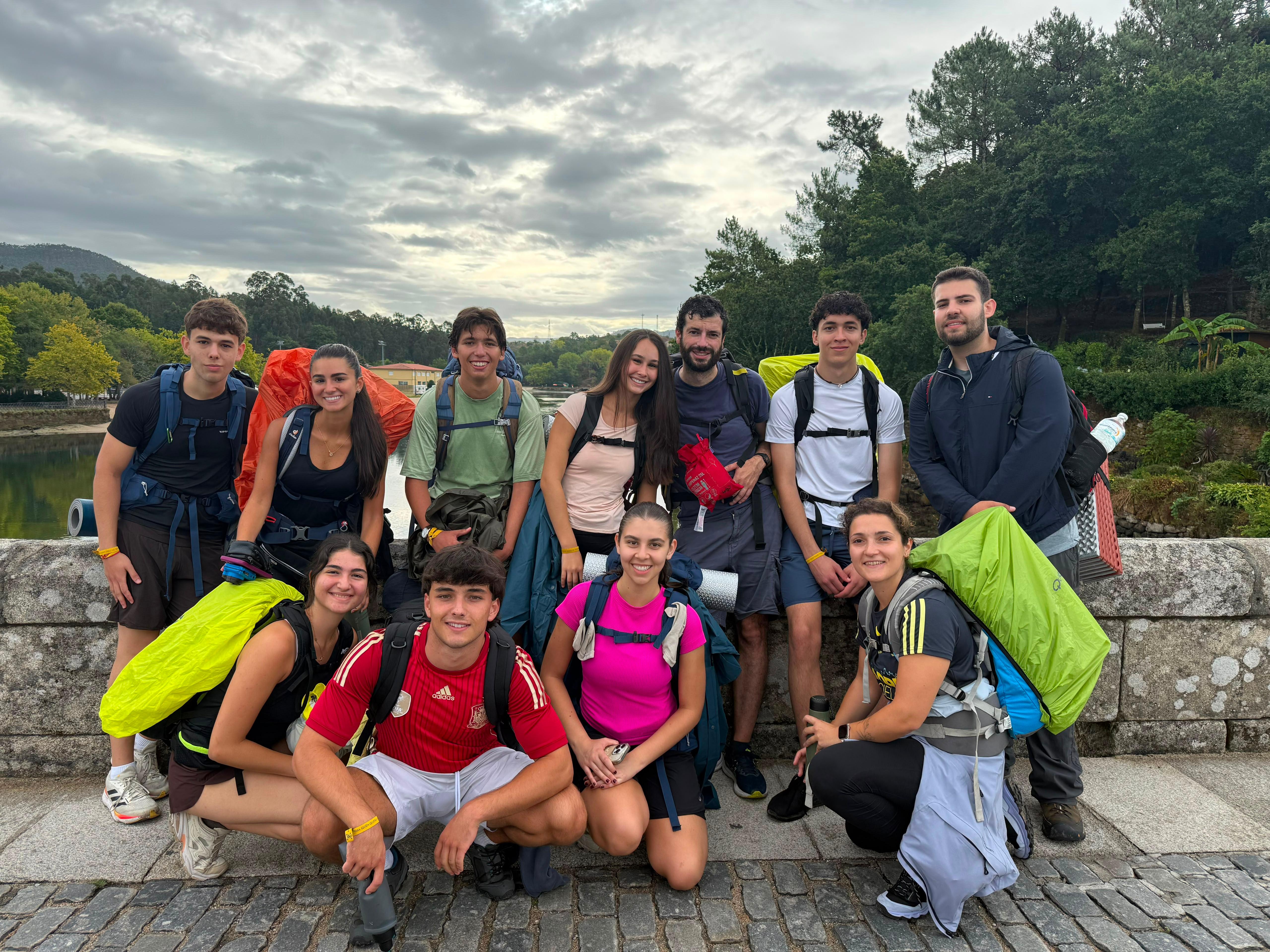 Grupo de jóvenes posando en un puente con mochilas, rodeados de naturaleza y un paisaje de fondo.