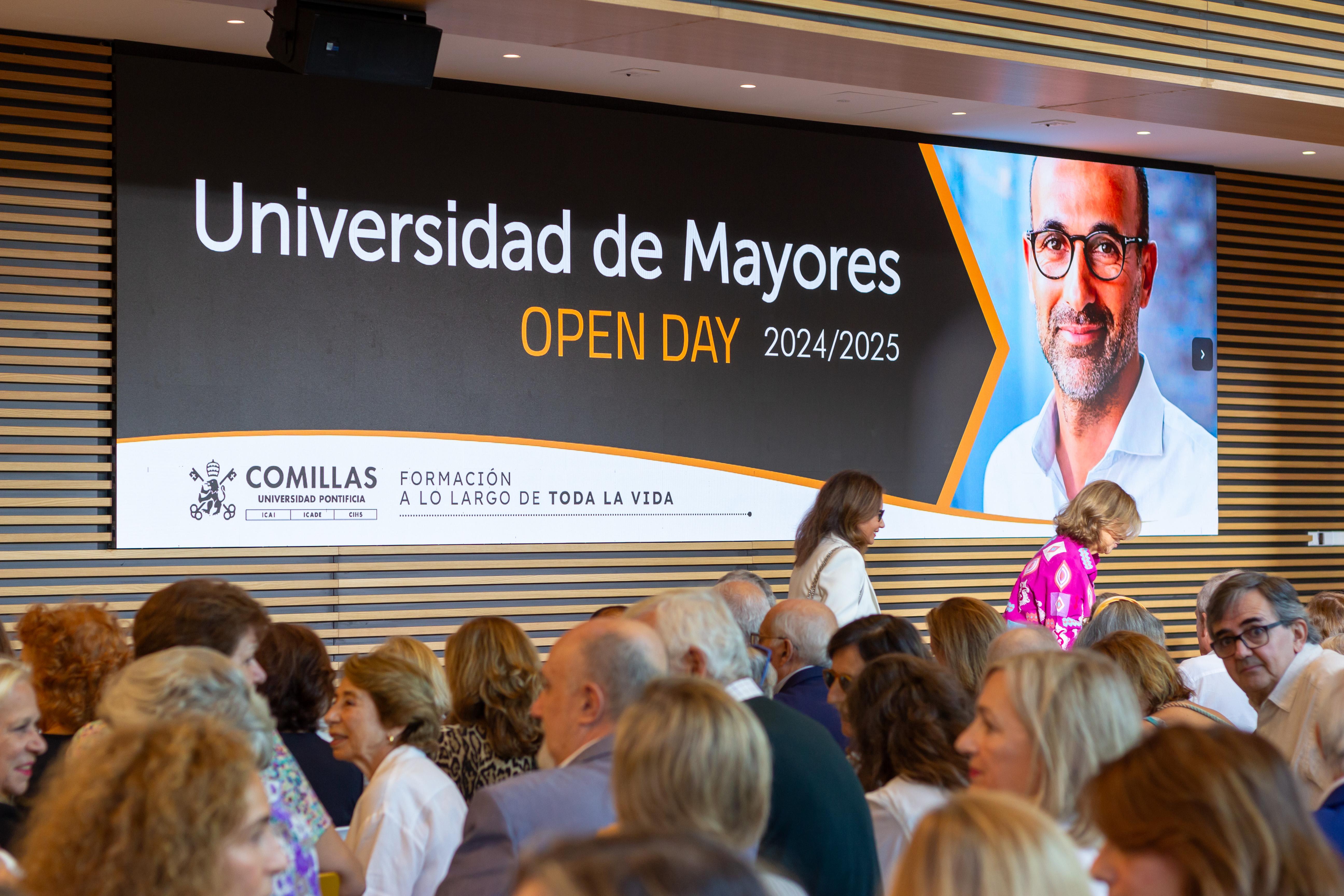 A gathering of people at an open day event for Universidad de Mayores, with a large digital banner featuring a man's portrait in the background.