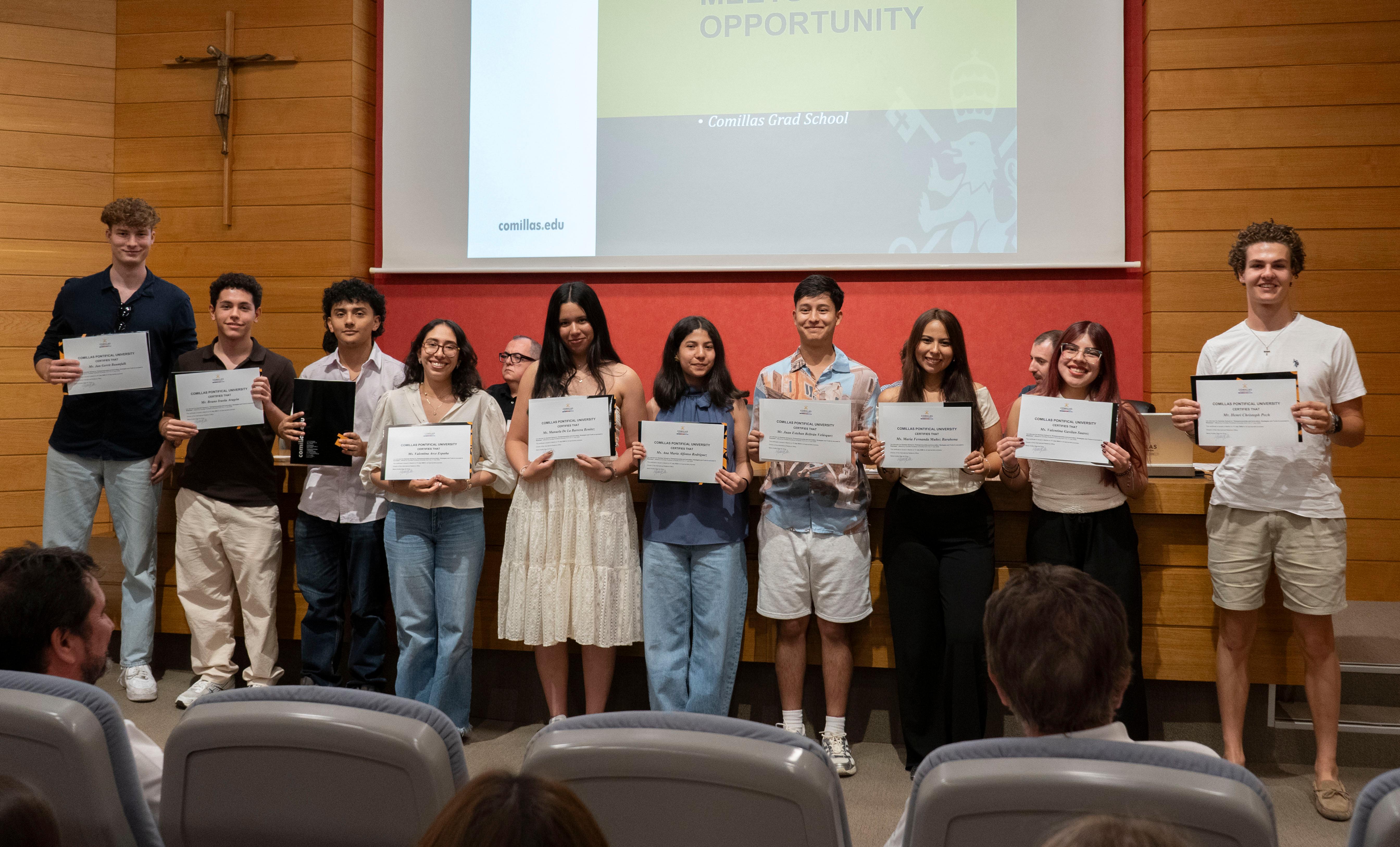 Un grupo de estudiantes sonrientes se encuentra en un escenario, mostrando sus certificados de logro frente a una audiencia.