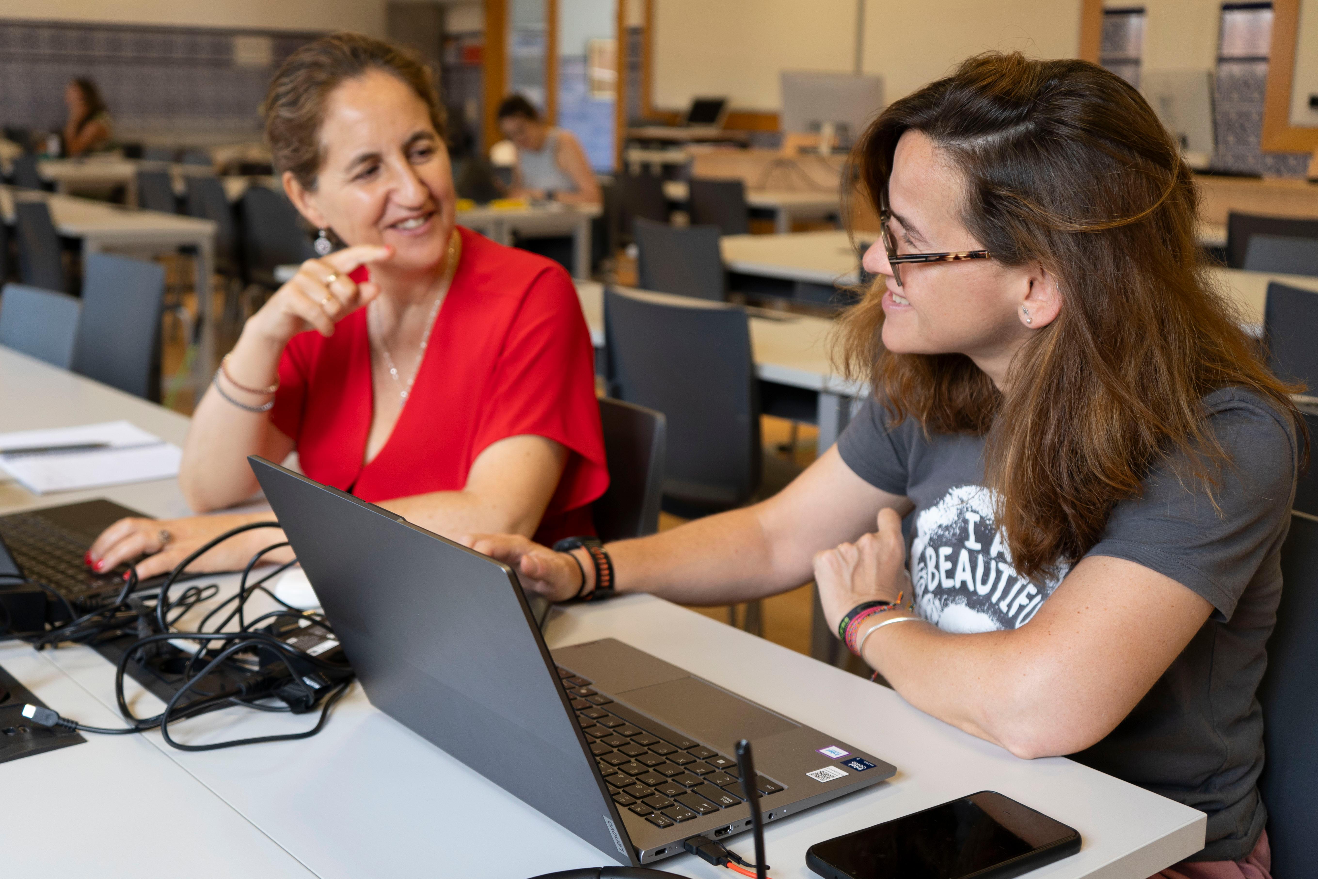 Dos mujeres están conversando y trabajando en una computadora portátil en un espacio de estudio.