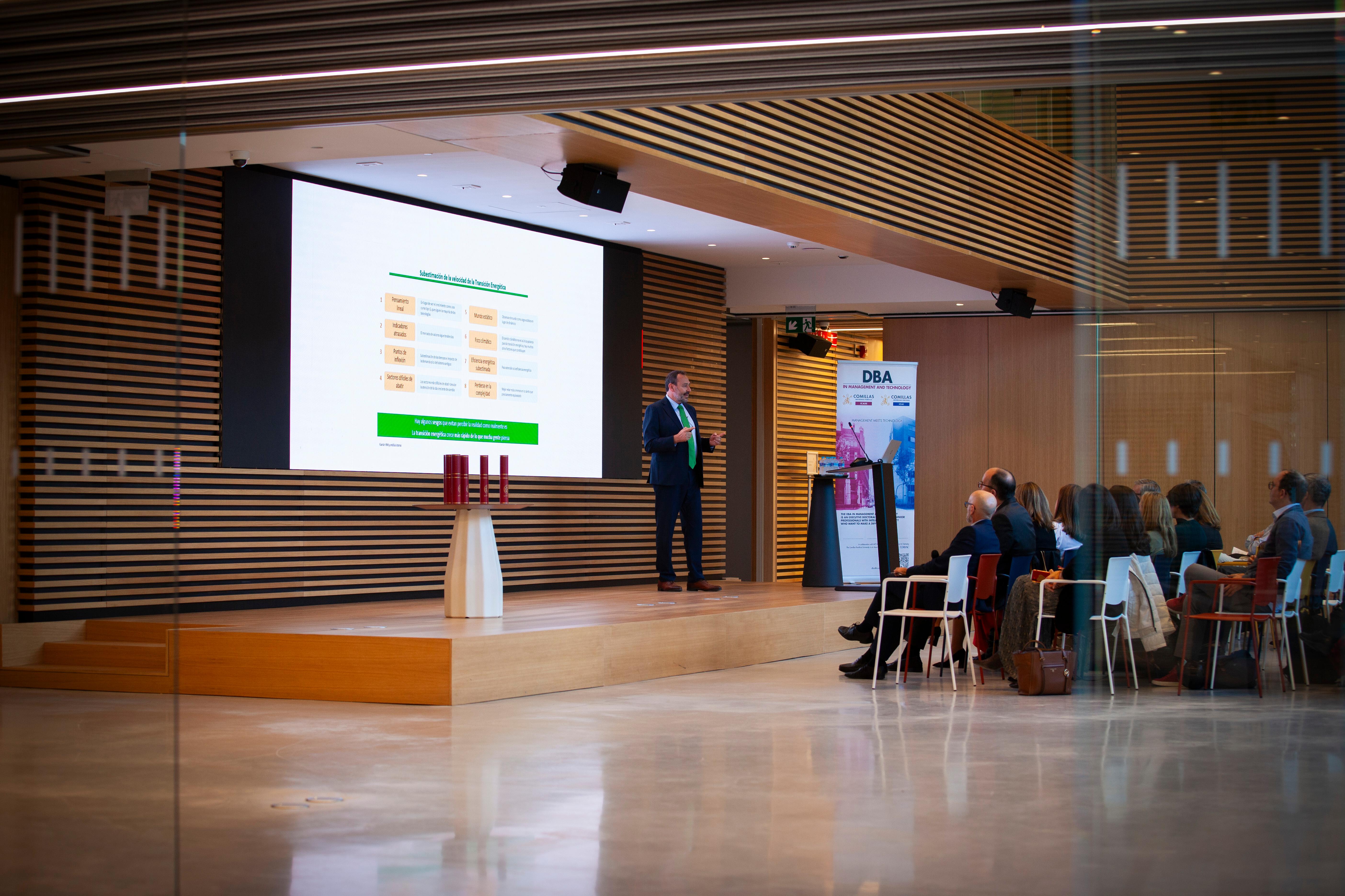 A man presenting at a conference in a modern hall with attendees seated and watching.