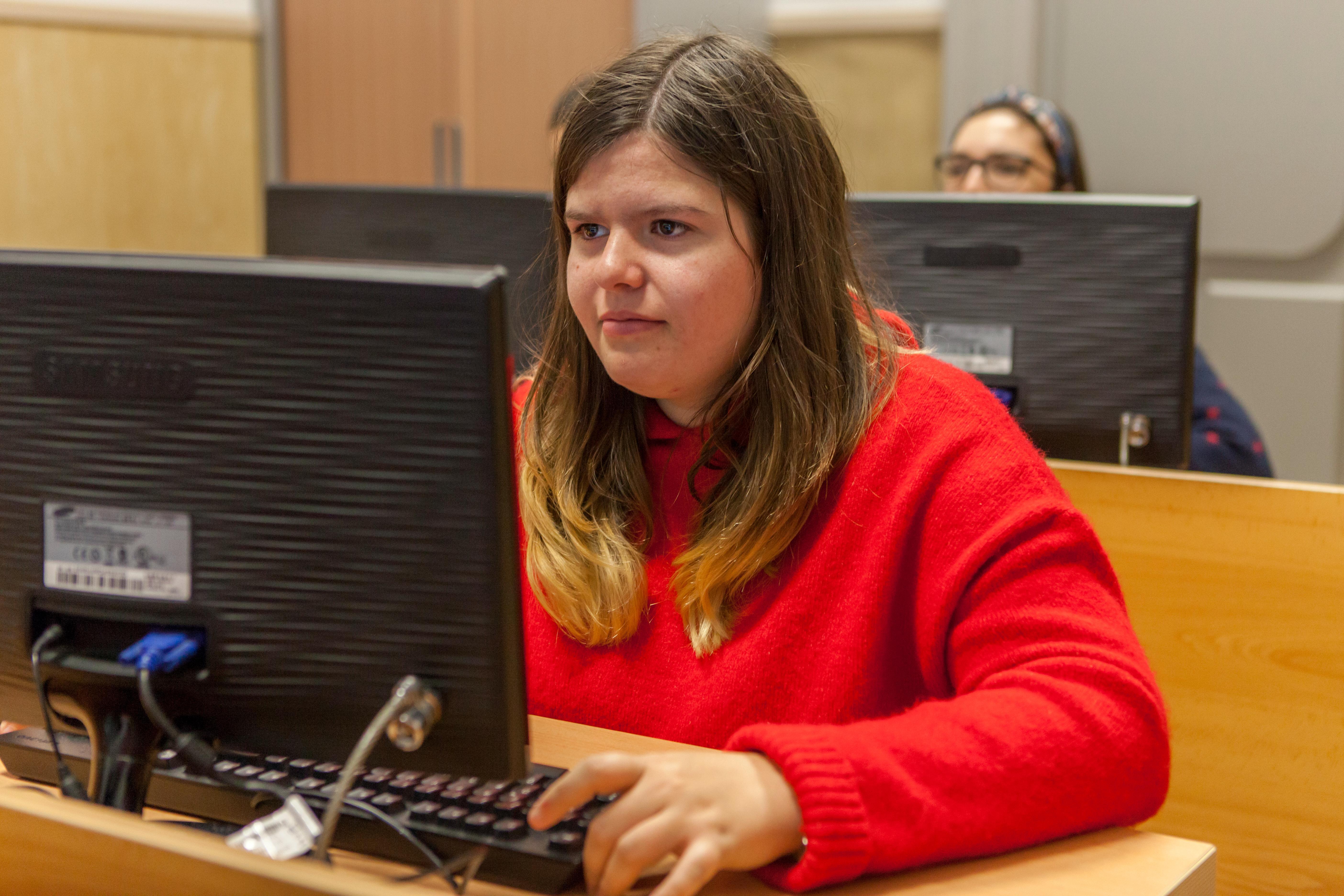 A woman in a red sweater is focused on her computer in a classroom setting.