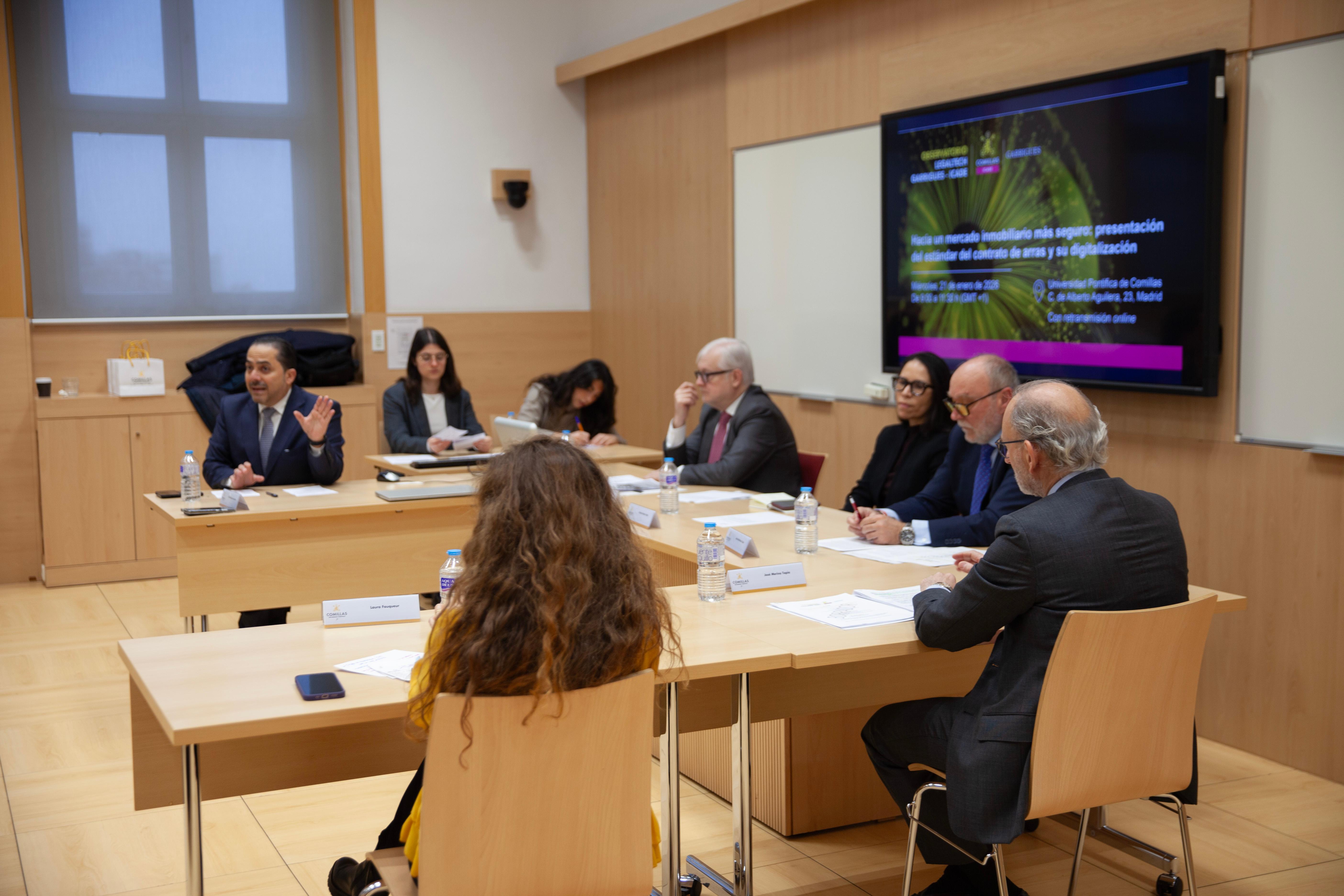 Una reunión en una sala con varias personas sentadas alrededor de una mesa discutiendo un tema presentado en una pantalla.