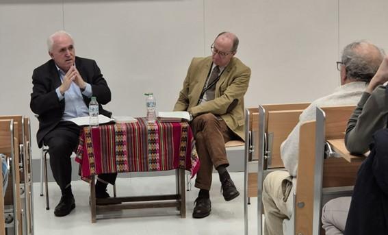 Tres hombres sentados en sillas frente a un público durante una discusión o conferencia en un espacio cerrado.