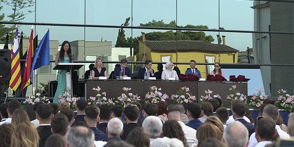 A woman is giving a speech at an outdoor graduation ceremony, with several people seated on stage and an audience in front.