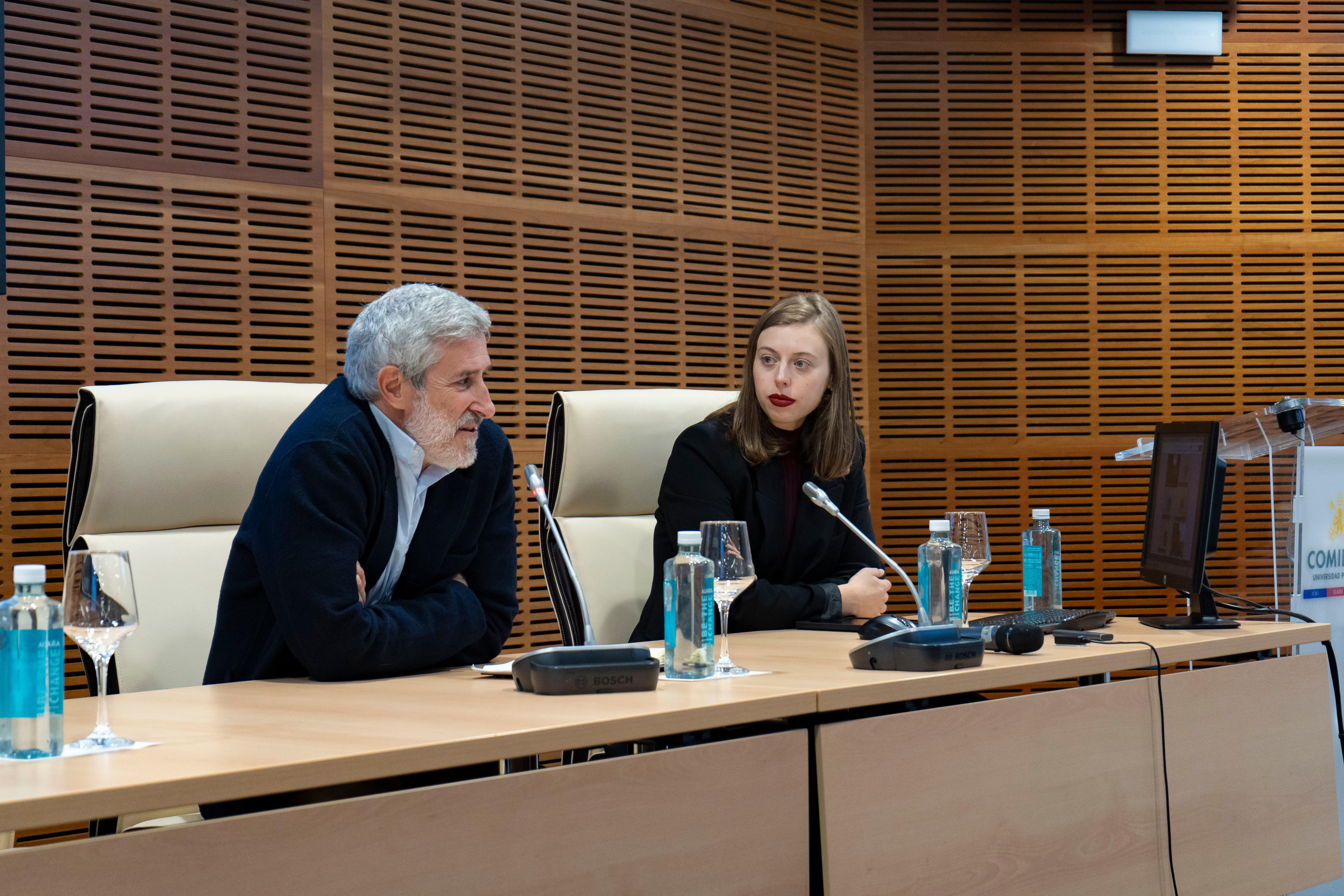 Un hombre y una mujer sentados en una mesa de conferencias, con micrófonos delante y un fondo de pared de madera.