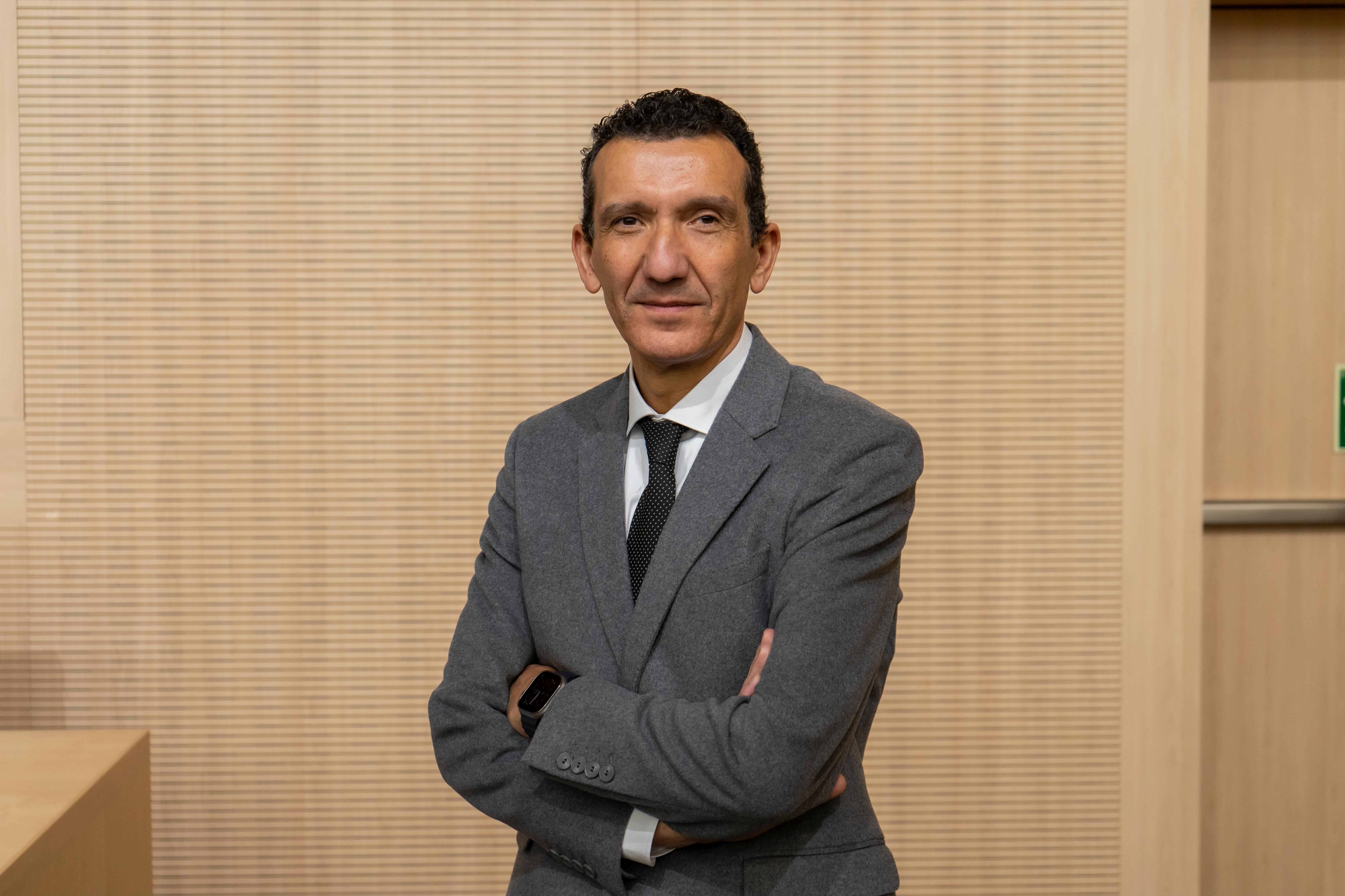 Un hombre de pie con brazos cruzados, vestido con un traje gris y una corbata negra, frente a un fondo de madera claro.