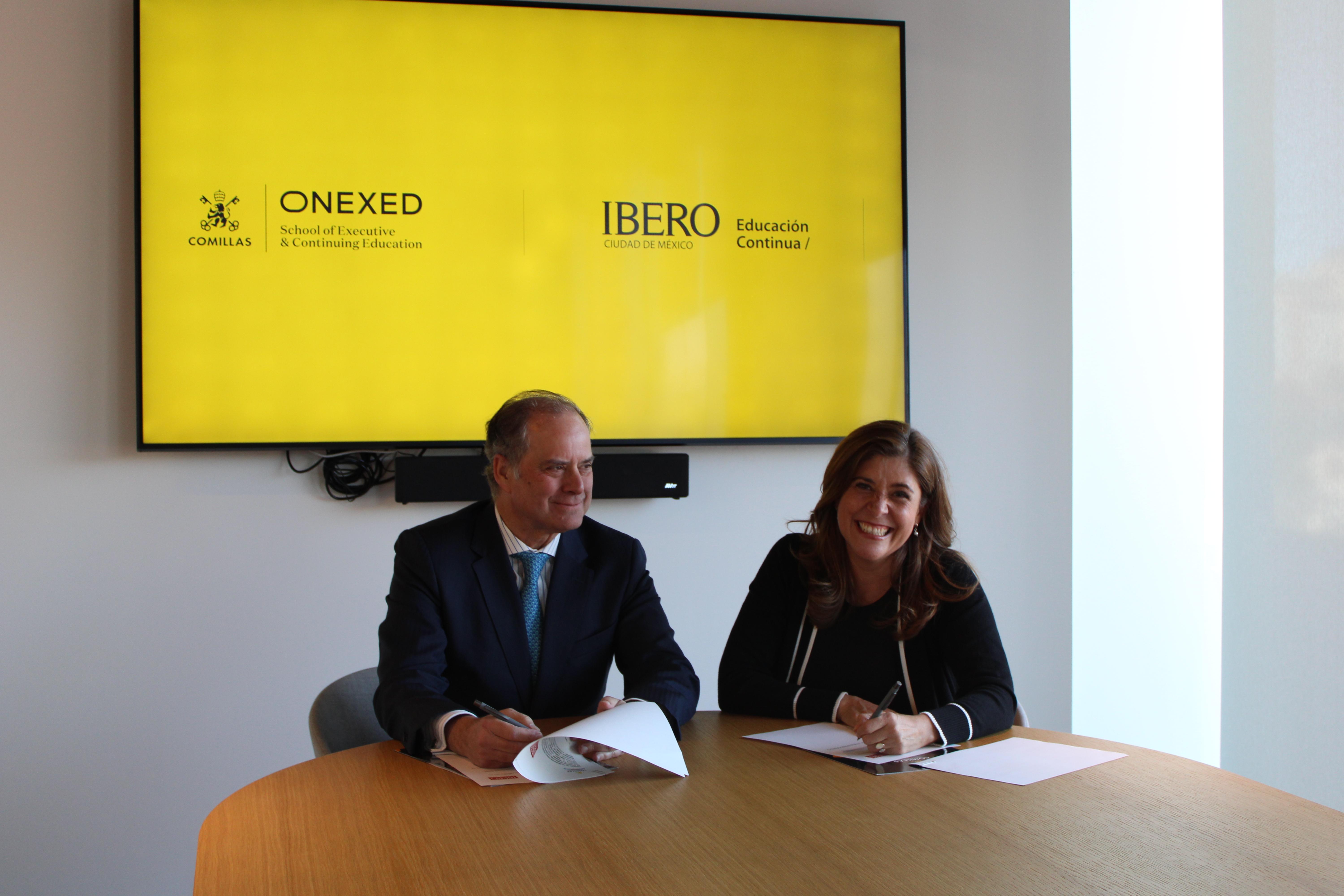 Dos personas sentadas en una mesa firmando documentos con una pantalla de fondo que muestra logotipos de ONEXED e IBERO.