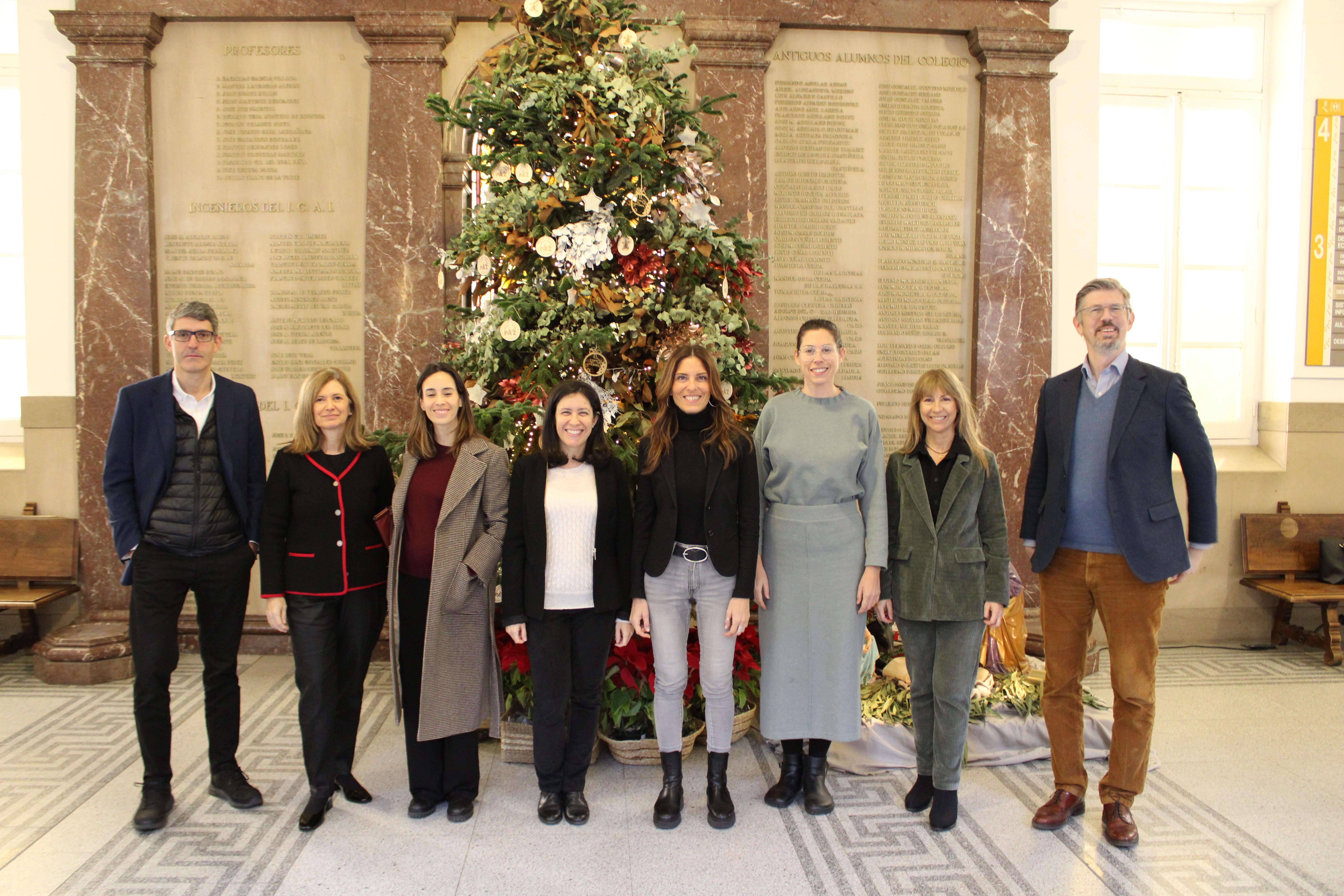 Un grupo de siete personas posando sonriendo frente a un gran árbol de Navidad decorado, en un interior elegante con columnas.