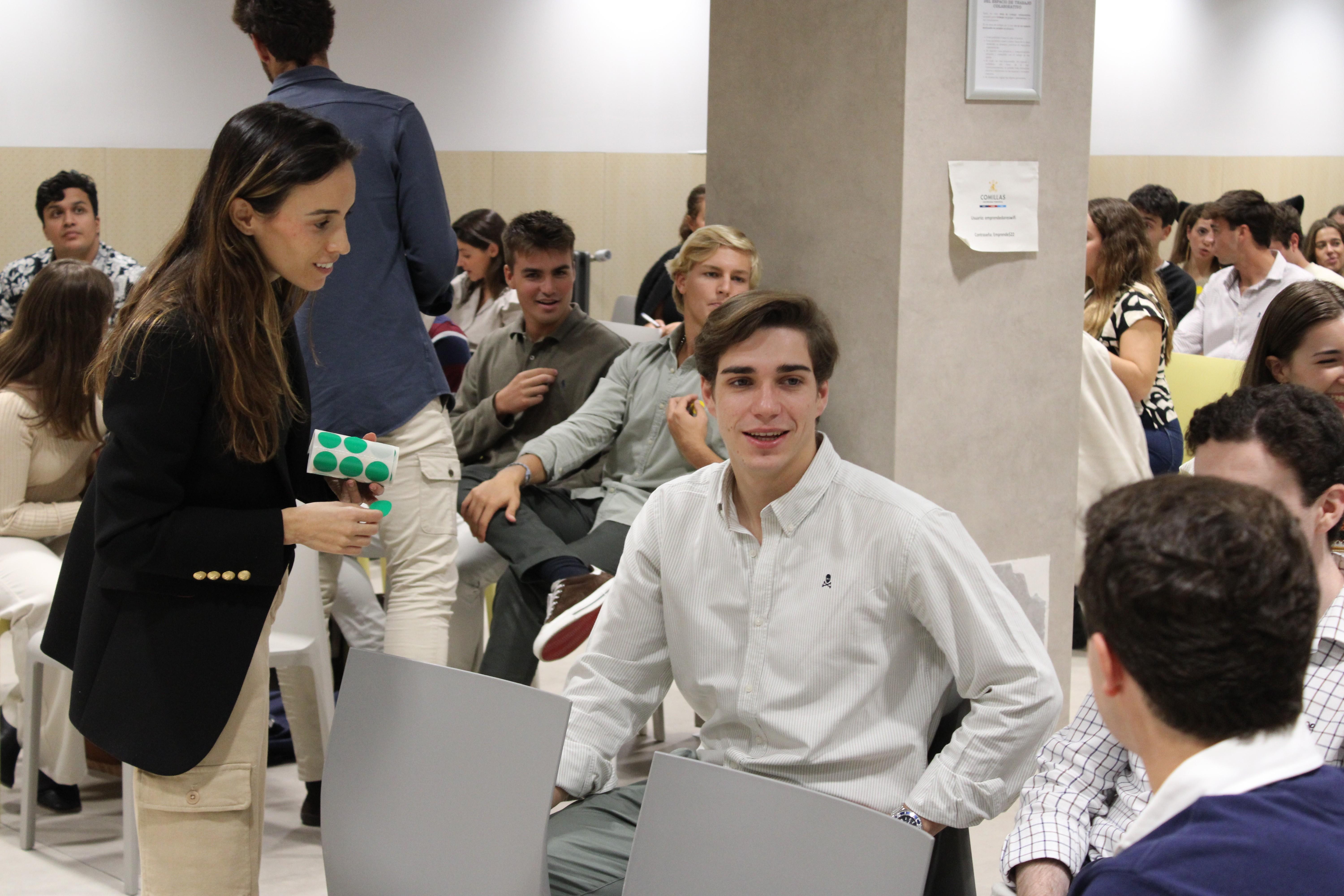 A group of young adults is seated in a modern classroom, engaging with each other and using laptops.