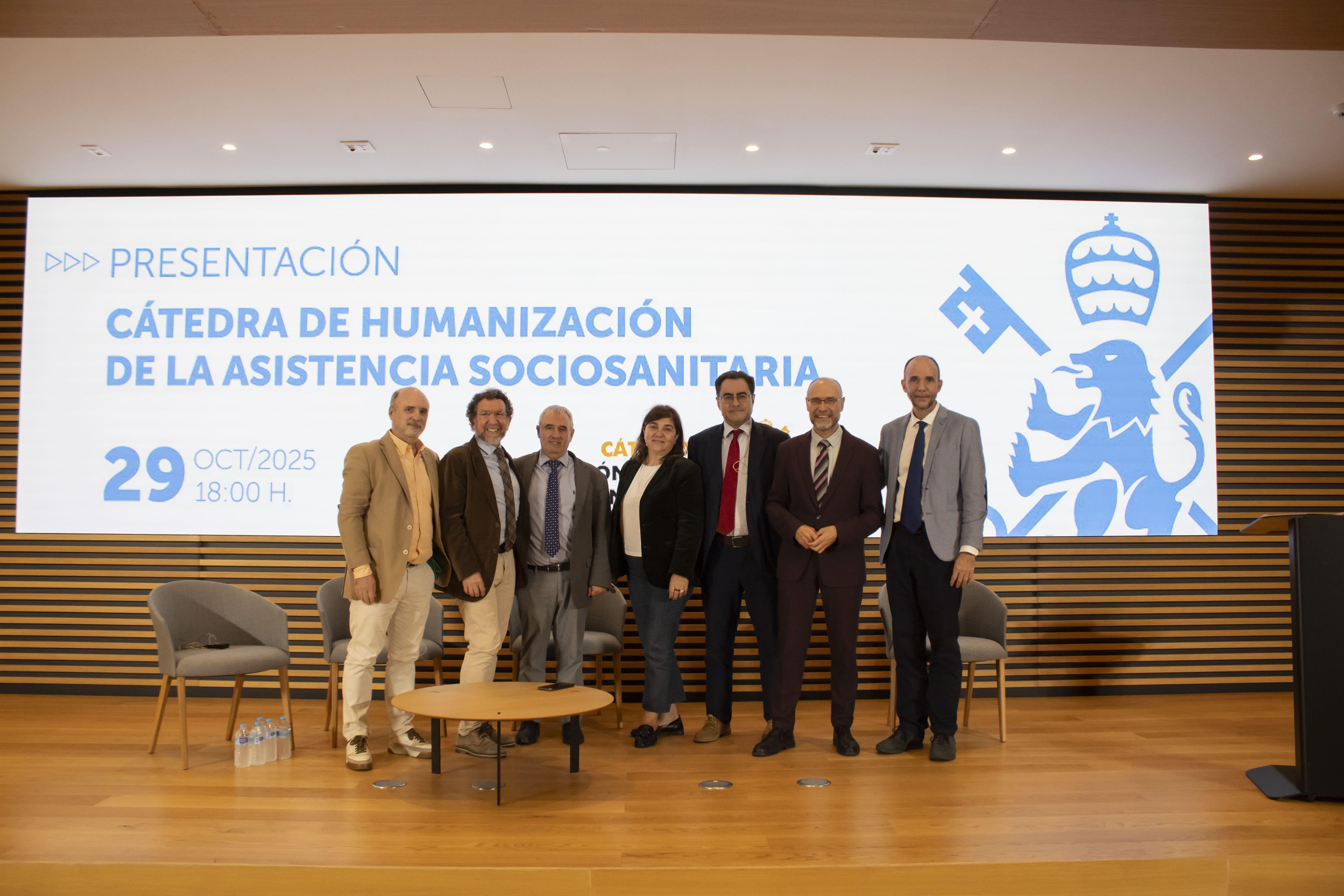 Un grupo de personas posando frente a un cartel sobre la presentación de la Cátedra de Humanización de la Asistencia Sociosanitaria.