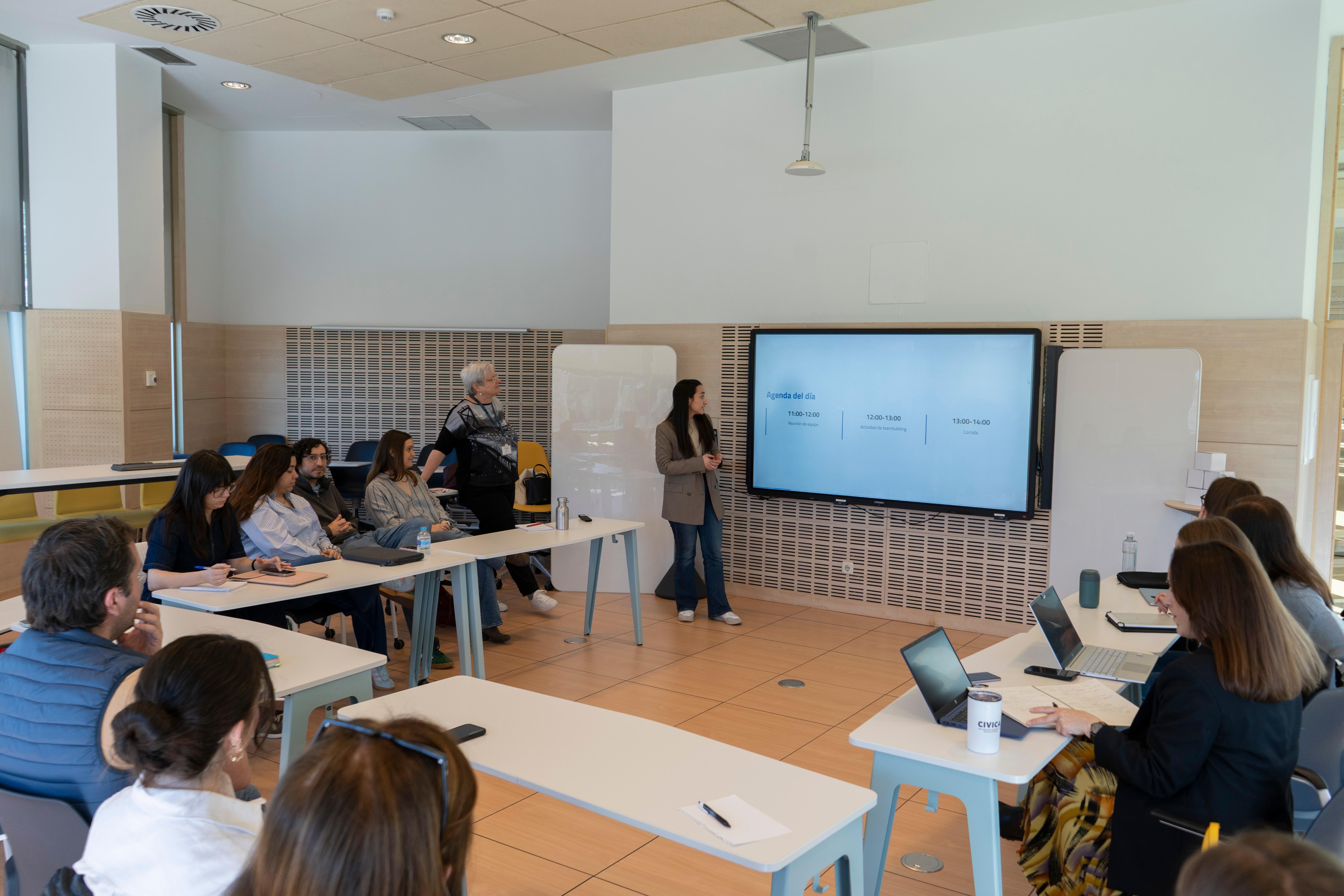 Una presentación en un aula con varias personas sentadas y una expositora en la pantalla.
